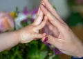 Mão de criança encostando palma na mão de adulto diante de flores desfocadas, representando afeto materno