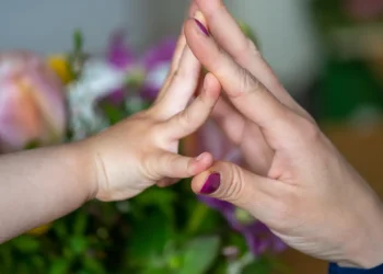 Mão de criança encostando palma na mão de adulto diante de flores desfocadas, representando afeto materno