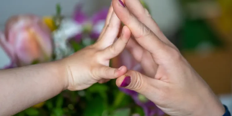 Mão de criança encostando palma na mão de adulto diante de flores desfocadas, representando afeto materno