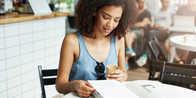 Jovem estudante lendo material de estudo em cafeteria enquanto segura um copo de café