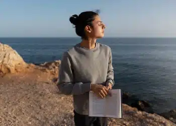 Mulher em momento de introspecção à beira-mar segurando um livro aberto durante o pôr do sol.