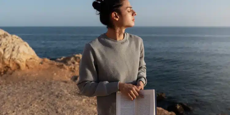 Mulher em momento de introspecção à beira-mar segurando um livro aberto durante o pôr do sol.