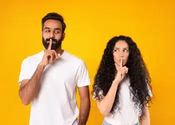 Homem e mulher fazendo gesto de silêncio com o dedo nos lábios, representando a importância de manter informações pessoais em sigilo.