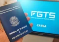 Mão segurando carteira de trabalho azul em frente a monitor exibindo logo do FGTS e Caixa
