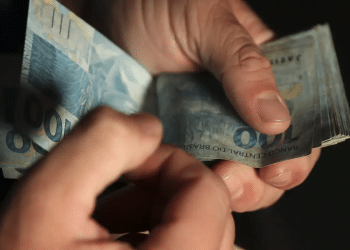 Mãos humanas contando notas de cem reais em ambiente escuro, simbolizando a contagem dos pagamentos do seguro-defeso 2026.