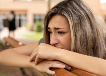 Mulher jovem com expressão triste e pensativa, apoiando o rosto sobre as mãos em um banco de madeira ao ar livre, representando o peso emocional da culpa.