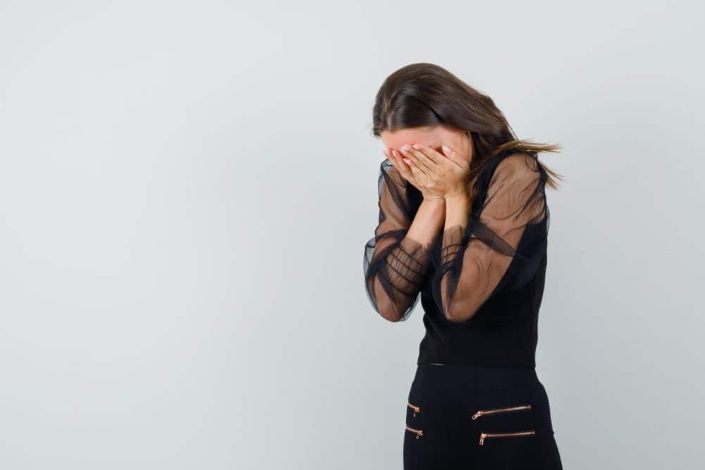 Uma mulher jovem de perfil, usando blusa preta de tule transparente, cobre o rosto com as mãos em um gesto de angústia ou retração social contra um fundo cinza claro.