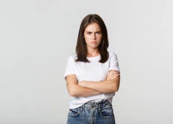 Uma mulher jovem de camiseta branca e calça jeans, com os braços cruzados e uma expressão facial de descontentamento e irritação contra um fundo branco.