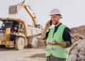 Homem jovem sorridente, com barba curta, usando capacete de segurança branco e colete refletivo verde neon sobre camisa cinza. Ele está falando ao celular em um canteiro de obras de mineração. Ao fundo, uma escavadeira amarela e um caminhão de carga pesada operam entre montes de terra e pedras sob luz solar clara.