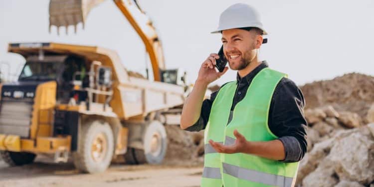 Homem jovem sorridente, com barba curta, usando capacete de segurança branco e colete refletivo verde neon sobre camisa cinza. Ele está falando ao celular em um canteiro de obras de mineração. Ao fundo, uma escavadeira amarela e um caminhão de carga pesada operam entre montes de terra e pedras sob luz solar clara.