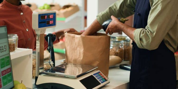 Funcionário pesando produto em balança digital no comércio, enquanto outro funcionário prepara sacola de papel, em contexto de atendimento ao cliente.