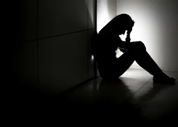 Pessoa sentada no chão, de costas para a parede, com a mão na cabeça em sala escura