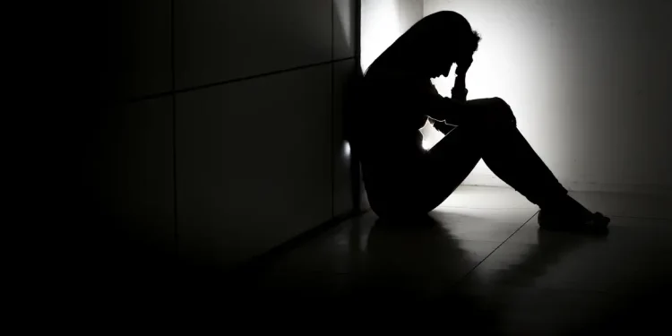 Pessoa sentada no chão, de costas para a parede, com a mão na cabeça em sala escura