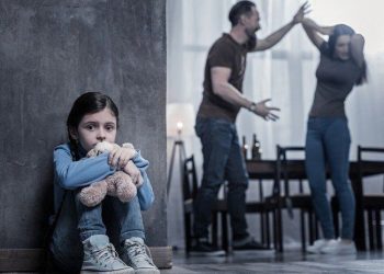 Criança sentada no chão abraçando ursinho enquanto casal discute ao fundo