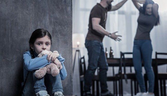 Criança sentada no chão abraçando ursinho enquanto casal discute ao fundo