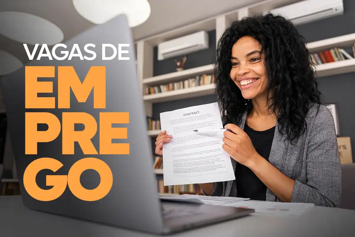 Mulher negra sorridente com cabelos cacheados, vestindo blazer cinza, segurando um documento de contrato em frente a um notebook. Ao lado esquerdo, o texto gráfico em destaque diz "VAGAS DE EMPREGO" em letras grandes brancas e laranjas.