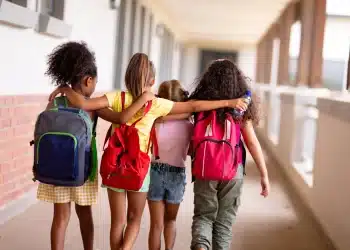 Grupo de crianças caminhando de mãos dadas em corredor escolar, com mochilas, simbolizando a volta às aulas em 2026.