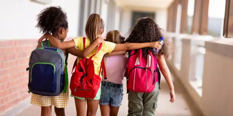 Grupo de crianças caminhando de mãos dadas em corredor escolar, com mochilas, simbolizando a volta às aulas em 2026.