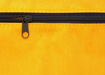 Close-up macro de um zíper preto com cursor metálico fechado em um tecido de lona amarela vibrante com costura visível.