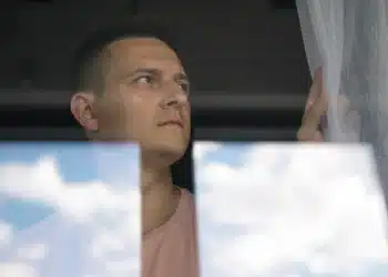 Homem olhando seriamente pela janela com cortina branca ao lado e reflexo de nuvens