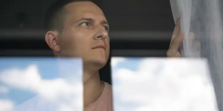 Homem olhando seriamente pela janela com cortina branca ao lado e reflexo de nuvens