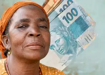 Mulher idosa com lenço laranja posa ao lado de várias notas de cem reais.