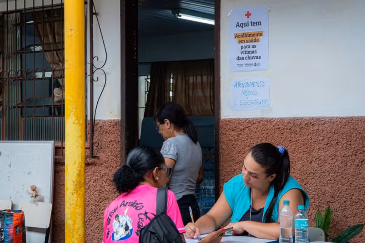 Profissional de saúde atende mulher sentada em mesa com papéis e garrafas de água.