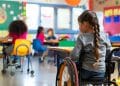 Criança com deficiência em cadeira de rodas participando de atividade em sala de aula inclusiva com outros estudantes.