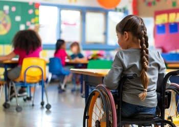 Criança com deficiência em cadeira de rodas participando de atividade em sala de aula inclusiva com outros estudantes.