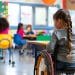 Criança com deficiência em cadeira de rodas participando de atividade em sala de aula inclusiva com outros estudantes.