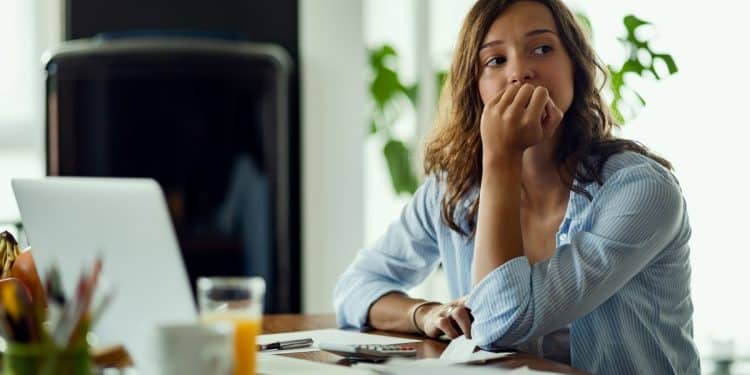 Mulher observa à distância apoiando a mão no rosto enquanto analisa documentos na mesa junto a notebook.