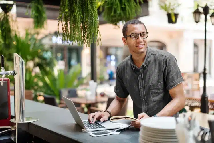 Jovem usando notebook e segurando caderno em cafeteria ao ar livre, rodeado de plantas