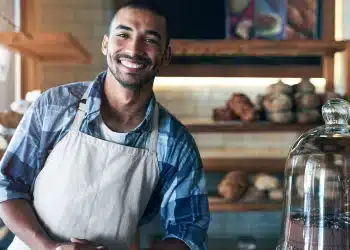 Homem jovem de avental sorri atrás do balcão com pães ao fundo e bolo em destaque