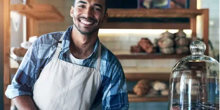 Homem jovem de avental sorri atrás do balcão com pães ao fundo e bolo em destaque
