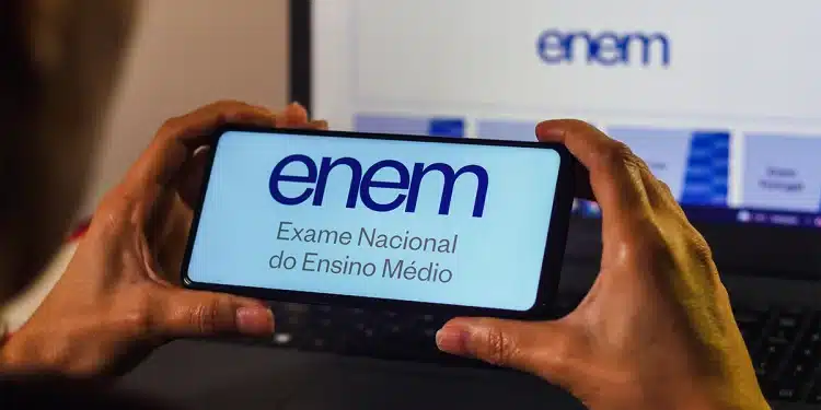 Pessoa segurando celular com a Página do Participante do Enem aberta enquanto acessa o sistema pelo notebook.