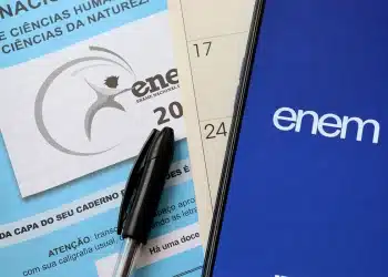 Foto de material do Enem com caderno de prova, caneta e celular exibindo o aplicativo oficial do exame.