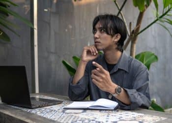 Homem jovem refletindo sobre ideias enquanto trabalha em seu laptop, praticando autodiálogo.