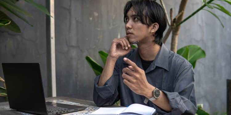 Homem jovem refletindo sobre ideias enquanto trabalha em seu laptop, praticando autodiálogo.