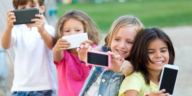 Quatro crianças tirando selfies com smartphones em parque, sorrindo e próximas umas das outras.