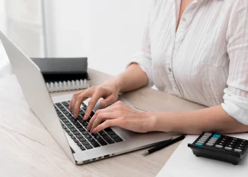 Mulher utilizando notebook ao lado de calculadora para verificar débitos e pendências fiscais com a nova ferramenta digital da Receita Federal.