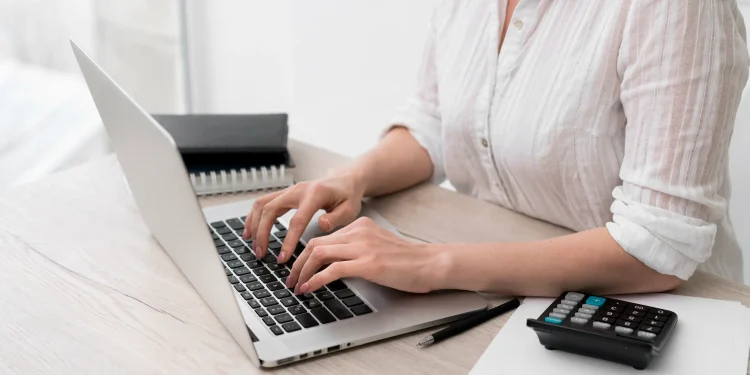 Mulher utilizando notebook ao lado de calculadora para verificar débitos e pendências fiscais com a nova ferramenta digital da Receita Federal.