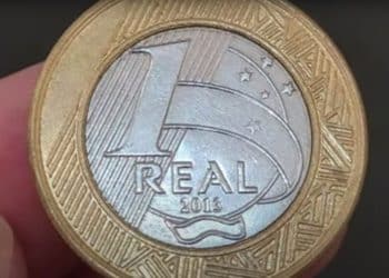 Close-up de uma moeda de 1 real de 2013 sendo segurada por dedos humanos, focando nos detalhes do valor "1 REAL", o ano de fabricação e as estrelas do Cruzeiro do Sul.