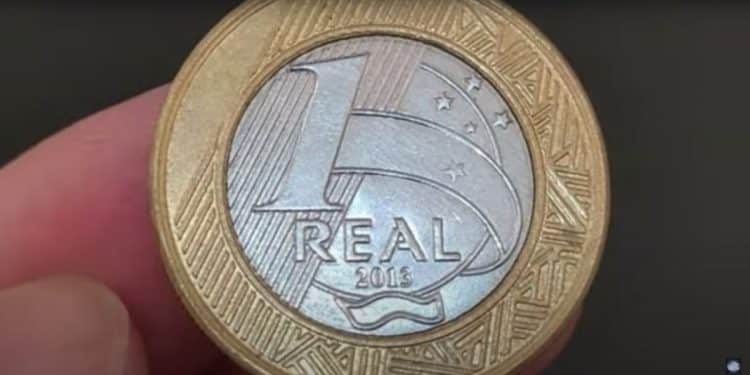 Close-up de uma moeda de 1 real de 2013 sendo segurada por dedos humanos, focando nos detalhes do valor "1 REAL", o ano de fabricação e as estrelas do Cruzeiro do Sul.