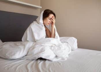 Jovem sentada na cama cobre-se com lençol branco enquanto segura a cabeça em sinal de desconforto.