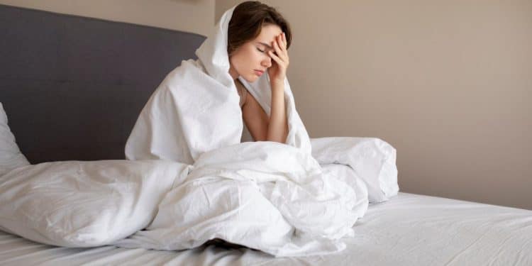 Jovem sentada na cama cobre-se com lençol branco enquanto segura a cabeça em sinal de desconforto.