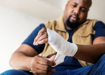 Homem ajustando bandagem na mão ferida com expressão de dor.