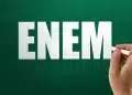 Mão escrevendo a palavra ENEM no quadro verde com giz branco, simbolizando o Exame Nacional do Ensino Médio.