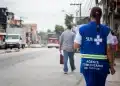 Profissional do SUS caminha em rua de bairro enquanto segura papéis, usando colete azul