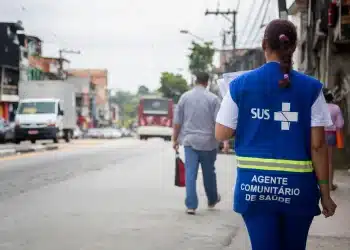 Profissional do SUS caminha em rua de bairro enquanto segura papéis, usando colete azul