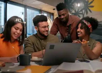 Quatro colegas de trabalho sorrindo e se comunicando durante uma reunião, demonstrando um ambiente de amizade e colaboração no escritório.